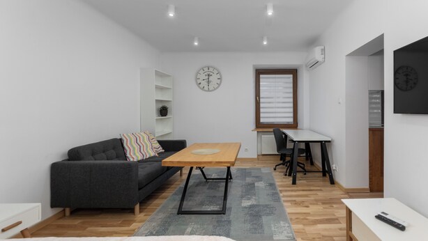 Habitació moderna i ben il·luminada amb un sofà gris, una taula de fusta i un escriptori petit. Hi ha un rellotge a la paret i una finestra amb persianes.