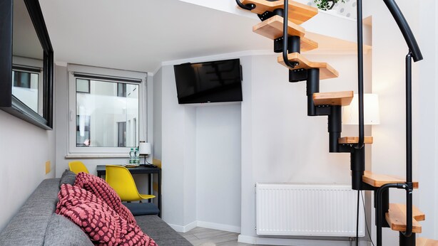 Una sala moderna con un sofá gris, un sillón amarillo, un plaid rojo estampado, un televisor negro en la pared y una escalera espiral de madera.