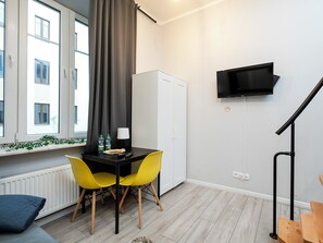 A modern room with a dining table, two yellow chairs, a white cabinet, a TV mounted on the wall, and a window with curtains. - Manufaktura Residence Studio #4B (Łódź)