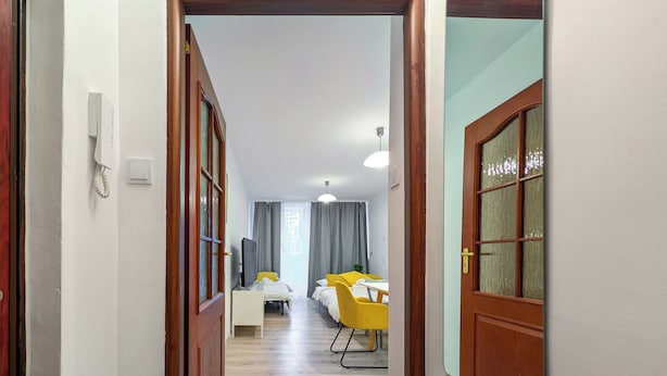Corredor que leva a um quarto com cama, escrivaninha e cadeira. Há um telefone e uma televisão na parede.