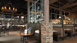 Een restaurantruimte met houten tafels en zithoekjes, een bar met flessenplanken, en een groot raam met het bordje 'SEVEN'.