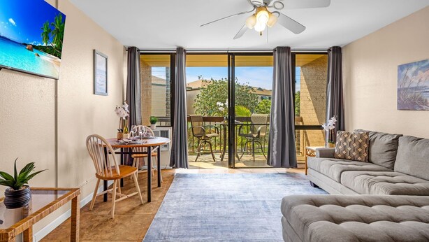 Ein Wohnzimmer mit einer Couch, einem Esstisch mit Stühlen, einem Deckenventilator und Blick durch eine Glastür auf einen Außenbereich mit Tisch und Stühlen.