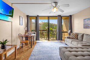 A living room with a sofa, a dining table with chairs, a ceiling fan, and a view through a glass door to an outdoor area with a table and chairs. - Bright and Chic Kihei Condo (Kihei)