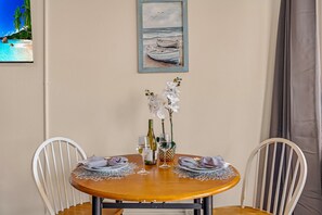 A round wooden table with white chairs, set for dining with plates, cutlery, and wine glasses. A painting of boats on water hangs above the table. - Bright and Chic Kihei Condo (Kihei)