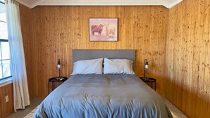 Un dormitorio con paredes de paneles de madera, una cama con ropa de cama gris, dos lámparas de noche y un cuadro enmarcado de toros en la pared.