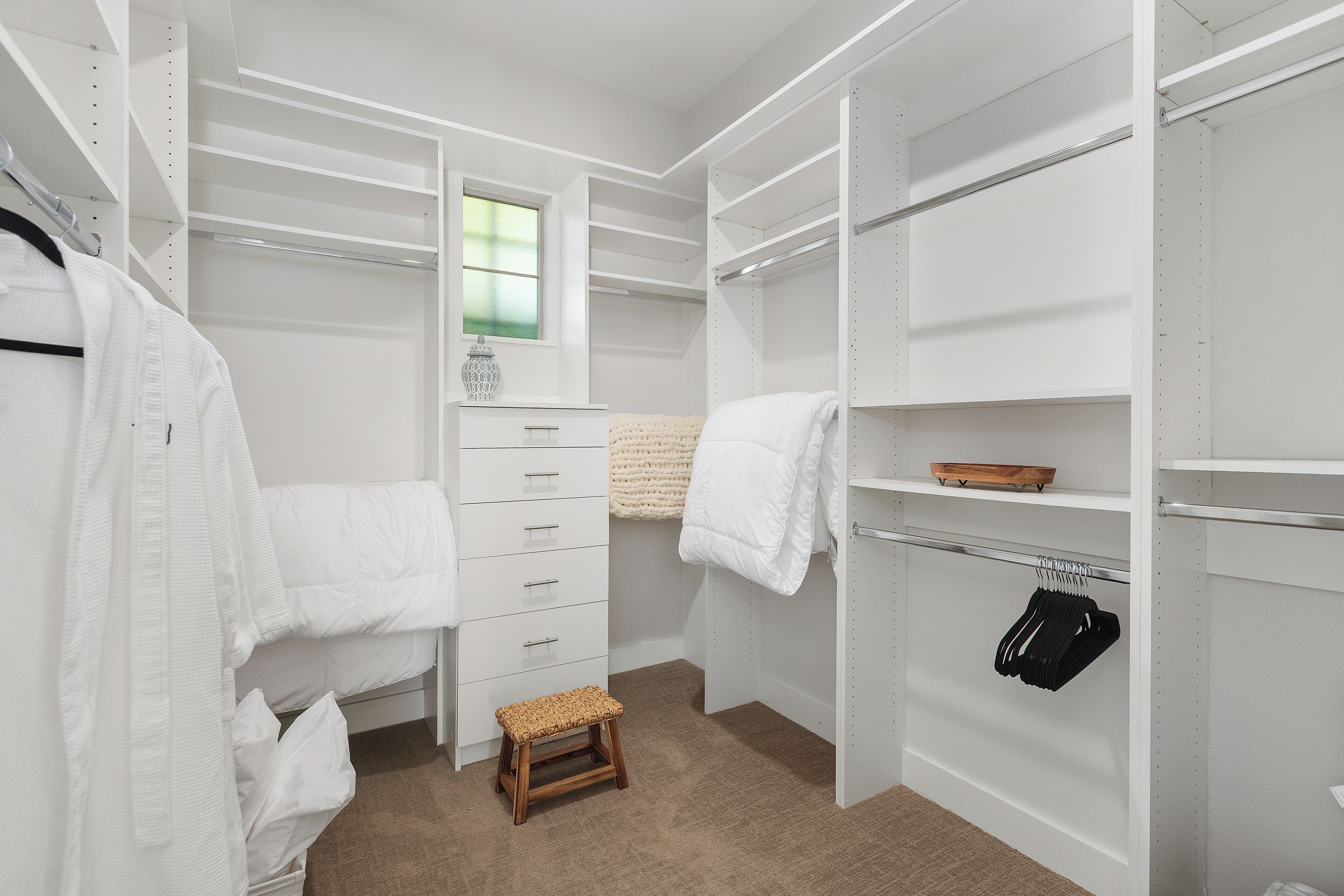 A walk-in closet with white shelving, a wooden stool, and a window.