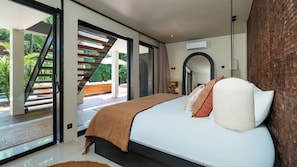 A bedroom with a large bed, a staircase leading to an outdoor area, and a view of greenery outside. - Villa Dreamora by Villa Finder (Seminyak)