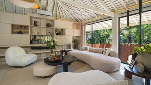 Een moderne woonkamer met een gebogen bank, een ronde salontafel en een witte fauteuil. Er zijn planken met decoratieve objecten en een groot raam met uitzicht op groen.