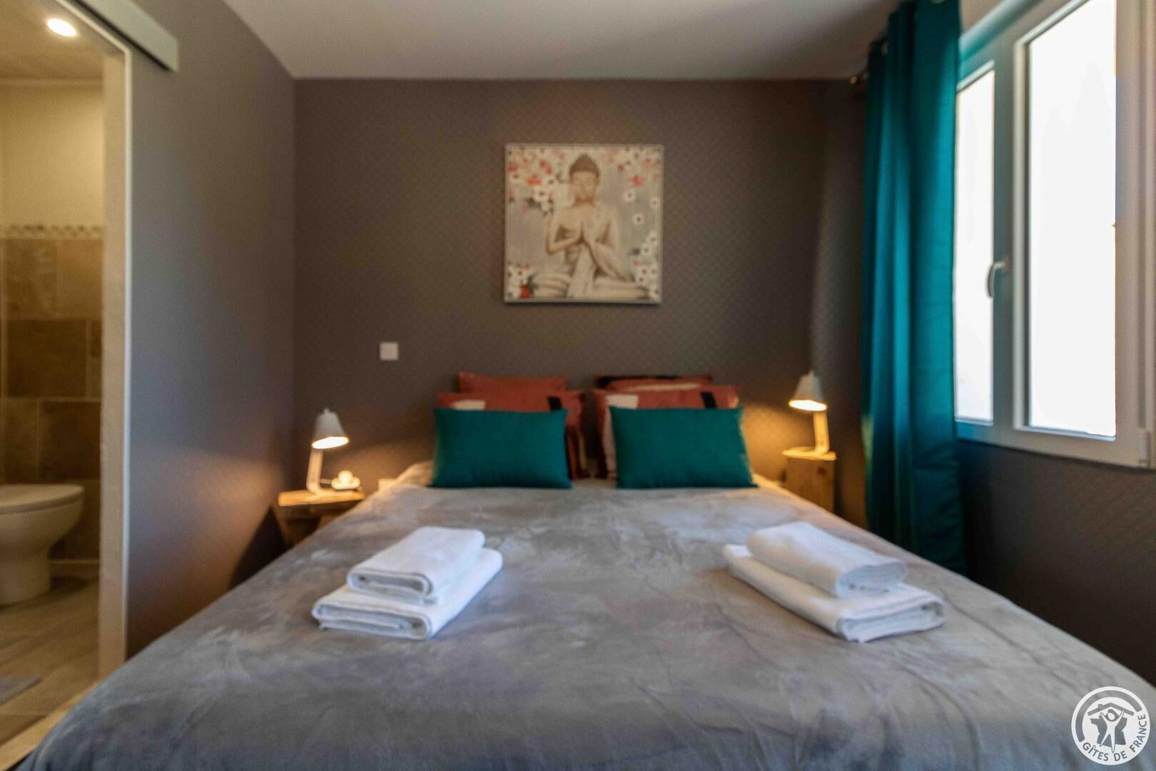 Hotelkamer met een bed, twee kussens, een nachtkastje met lamp, een schilderij aan de muur en een badkamer met toilet.