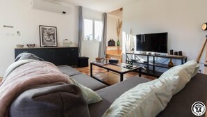 Ett modernt vardagsrum med en soffa, ett soffbord och en tv. Det finns ett fönster med gardiner, en bokhylla och en tavla på väggen.