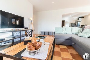 Ein modernes Wohnzimmer mit einem Flachbildfernseher, einem Couchtisch mit Gebäck, einem grauen Sofa und einer kleinen Küchenzeile im Hintergrund.