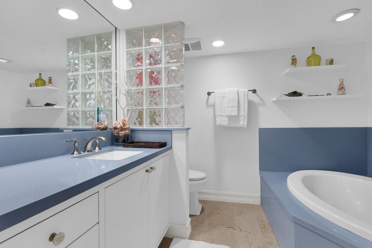 A modern bathroom with a large bathtub, a sink with a mirror, and a toilet. There are decorative items on shelves and a window with frosted glass panels.