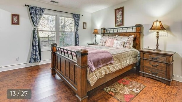 A bedroom with a wooden bed, a nightstand, a lamp, and a window with curtains. - Grill Patio • Pet Friendly • 3BR (Charlotte)