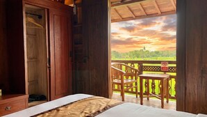 Una habitación de madera con cama, un armario y un balcón con vista a un atardecer sobre un paisaje verde.