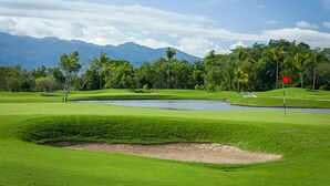 Ein Golfplatz mit Wasserhindernis, Palmen und Bergen im Hintergrund.