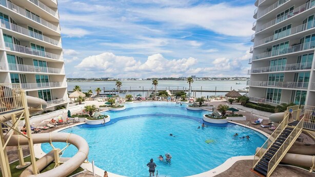 Une grande piscine intérieure avec un toboggan, entourée de chaises longues et de palmiers, et offrant une vue sur la mer et les bâtiments en arrière-plan.