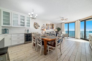 A spacious kitchen with white cabinets, a wooden dining table, and a large window offering a view of the sky. - Beachfront Escape | Gulf-View Balcony, Resort Pools & Iconic Location | BEACHBALL PROPERTIES (Orange Beach)