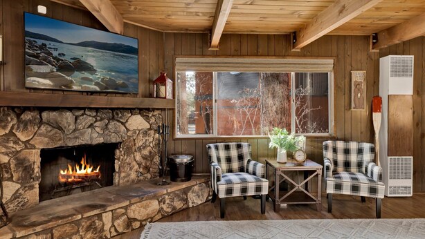 Interior acogedor de una cabaña con chimenea de piedra, sillones escoceses, una mesa con un reloj y un ventanal grande con vistas a un bosque.