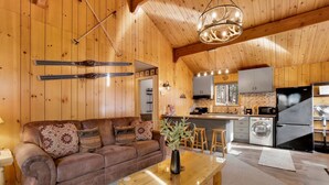 L’intérieur chaleureux d’un chalet, avec un revêtement mural en bois, un canapé, une table basse en verre et un coin cuisine équipé d’un réfrig érateur et d’un four à micro-ondes.