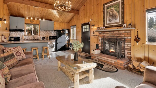 Un intérieur de chalet chaleureux, avec un revêtement en bois, une cheminée, un coin repas et une cuisine équipée d’appareils électroménagers modernes.