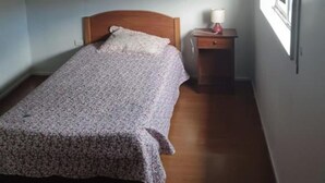 Une petite pièce, peu meublée, avec un lit simple, une table de chevet, une lampe et une fenêtre avec un rideau.