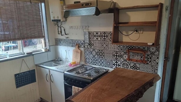 Keittiössä on kaasuliesi, valkoiset kaapit, kuvioidut laatat, ikkunassa sälekaihtimet ja puinen pöytä.