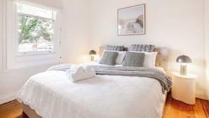 Een netjes opgemaakt bed met een witte deken, twee kussens en een opgevouwen handdoek. Daarboven hangt een lijst met een foto aan de muur, en ernaast staat een klein tafeltje met een lamp.