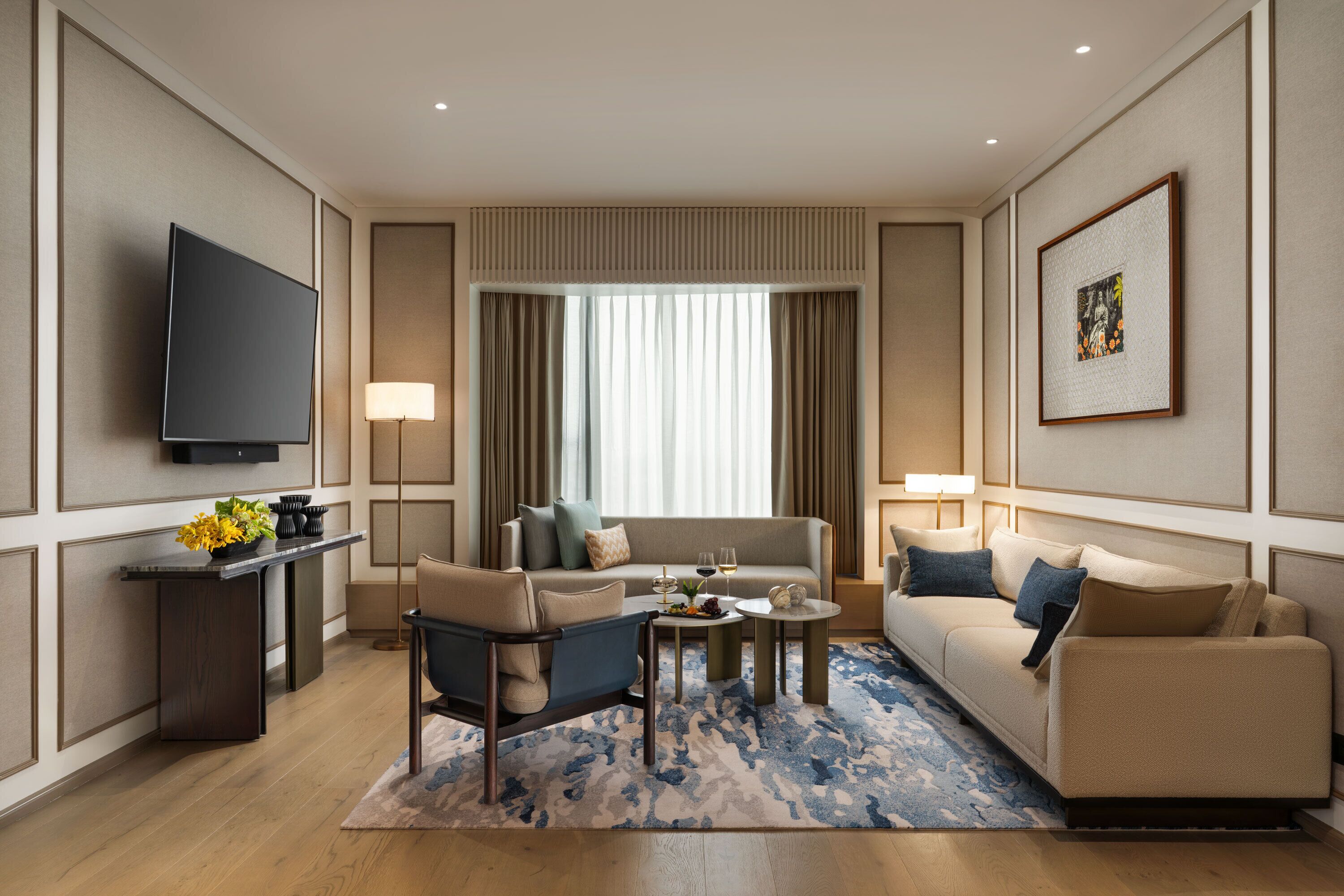 A modern hotel room with a flat-screen TV, a sofa, a chair, a coffee table, and a dining table with a vase of flowers.
