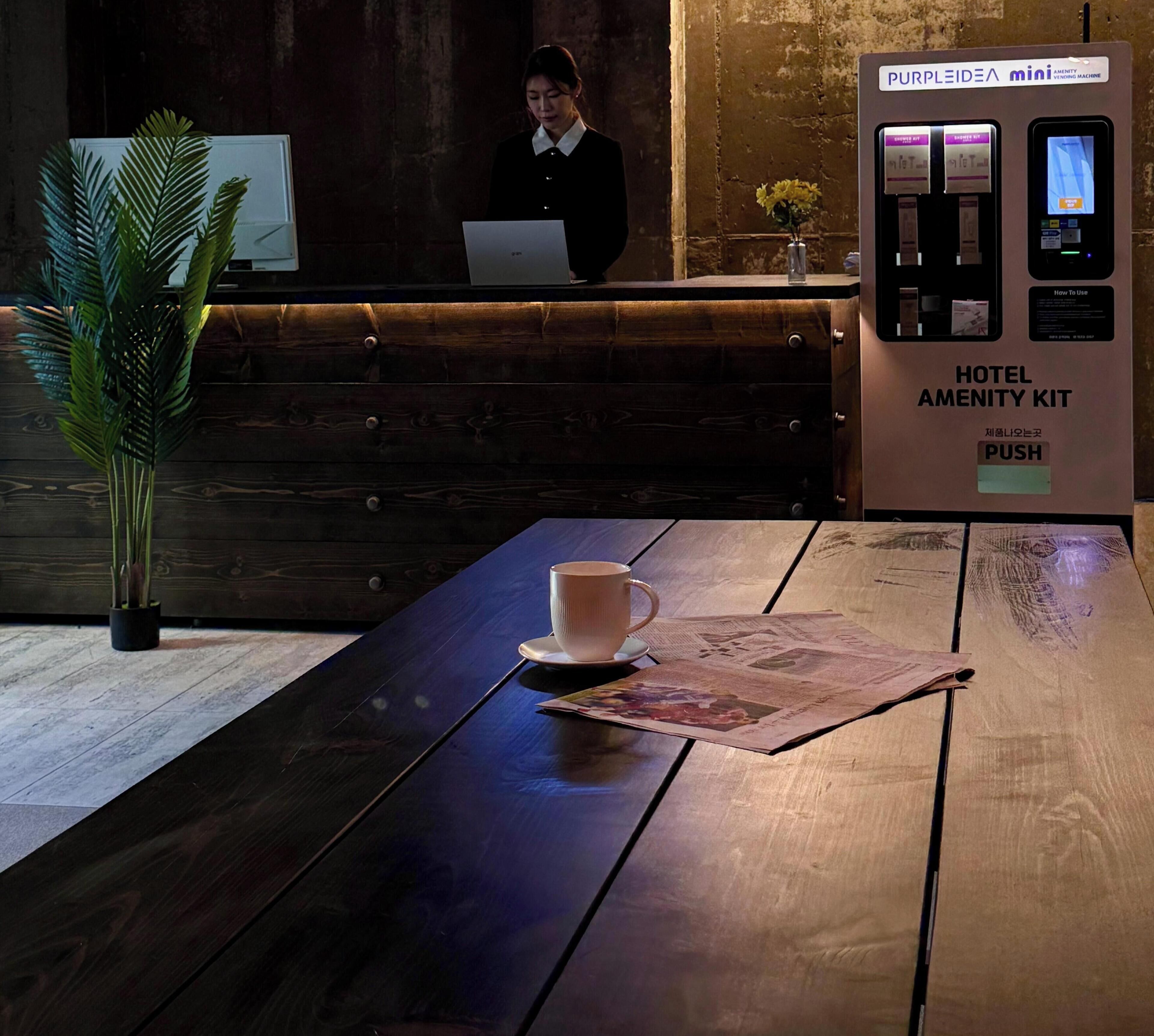 A hotel reception area with a wooden reception desk, a coffee cup on a coaster, a hotel amenity kit, and a purple vending machine.