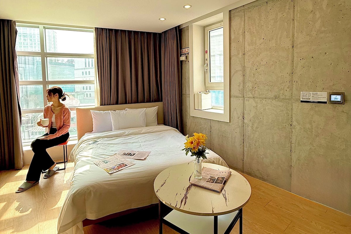 Hotelkamer met een bed, een klein tafeltje met een bloevas, een raam met gordijnen en uitzicht op gebouwen.