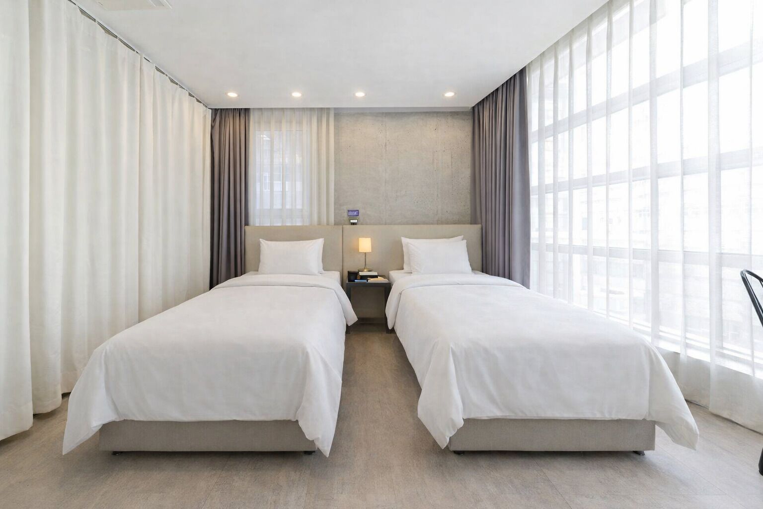 A hotel room with two beds, a nightstand, a lamp, and large windows with curtains.