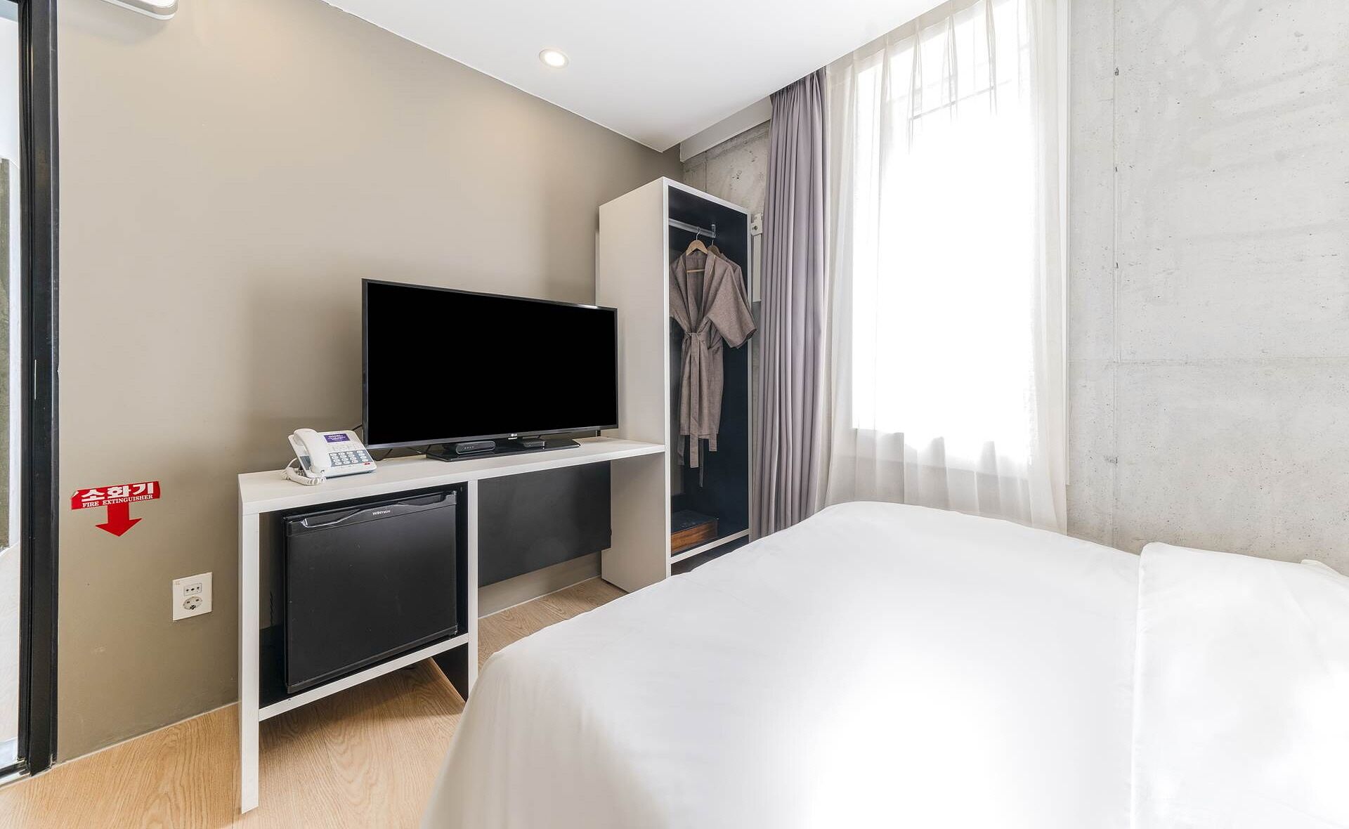 A hotel room with a bed, a TV on a stand, a wardrobe, and a fire extinguisher sign on the wall.