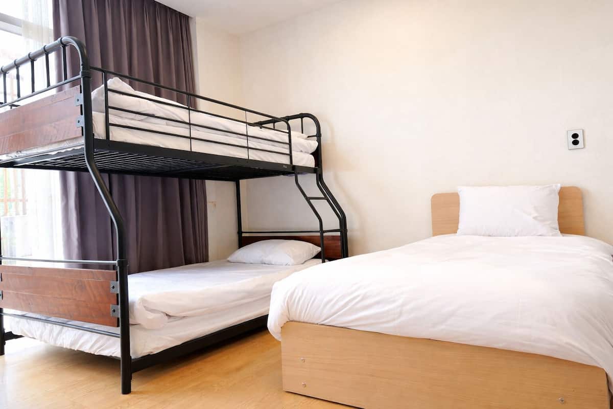 Een stapelbed met een onderbed en een eenpersoonsbed, beide met witte beddengoed, in een kamer met een houten vloer en een raam met gordijnen.