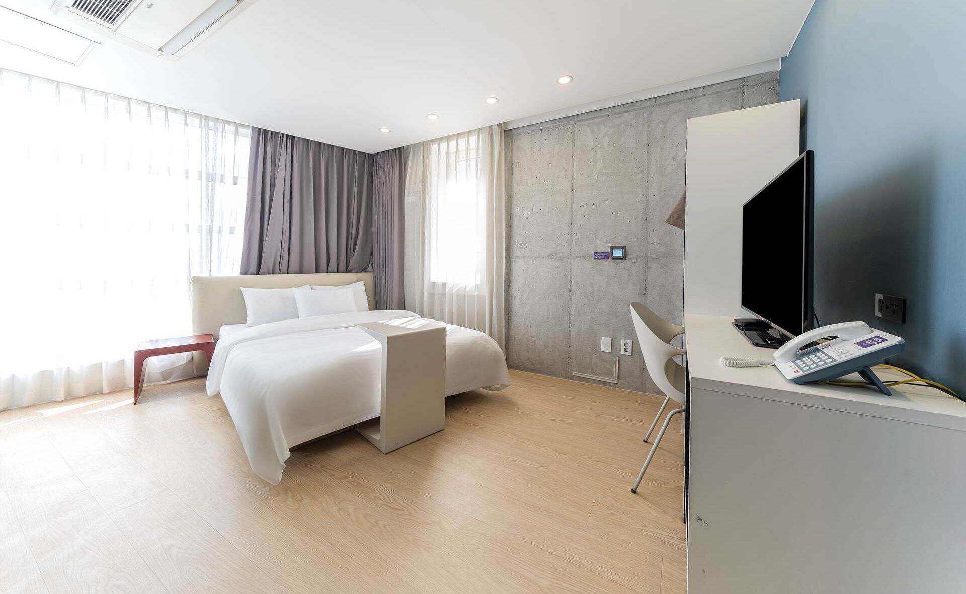 A modern hotel room with a bed, a desk with a phone and a flat-screen TV, a chair, and a large window with curtains.