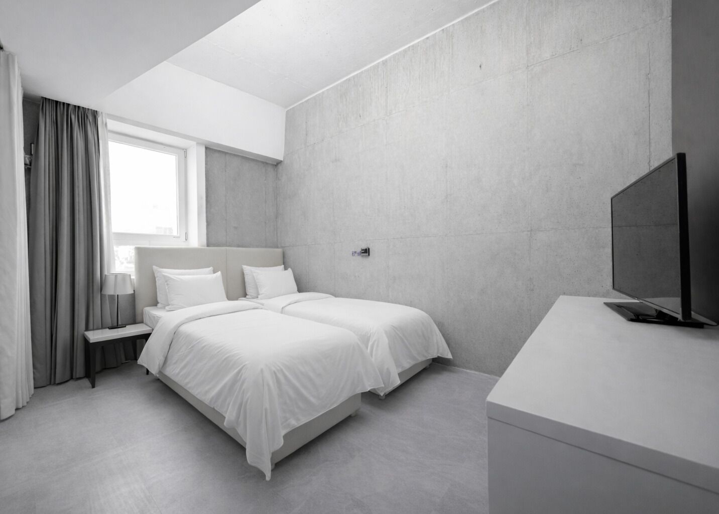 A modern hotel room with a large bed, a bedside table with a lamp, a flat-screen TV, and a large window with curtains.