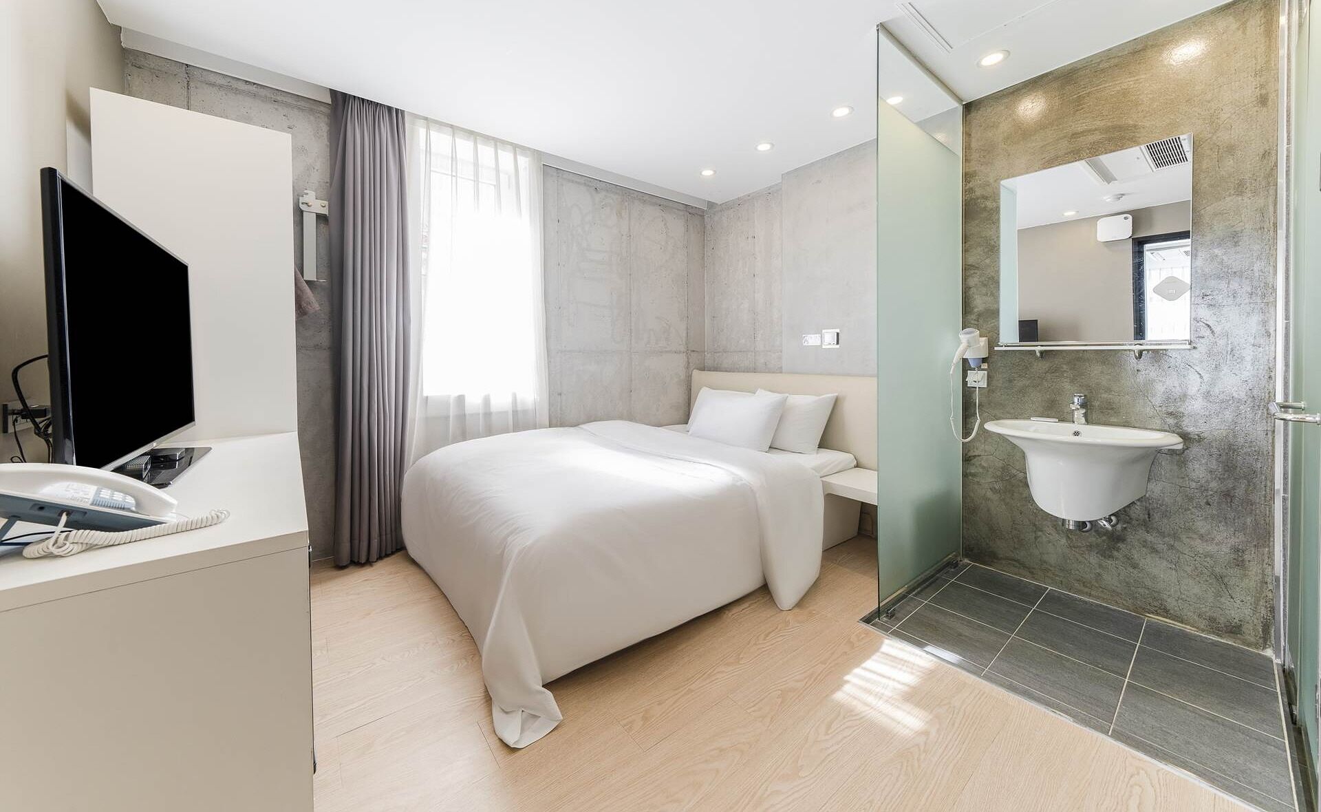A modern hotel room with a large bed, a bedside table with a phone, a TV, a bathroom with a sink and mirror, and a shower area.