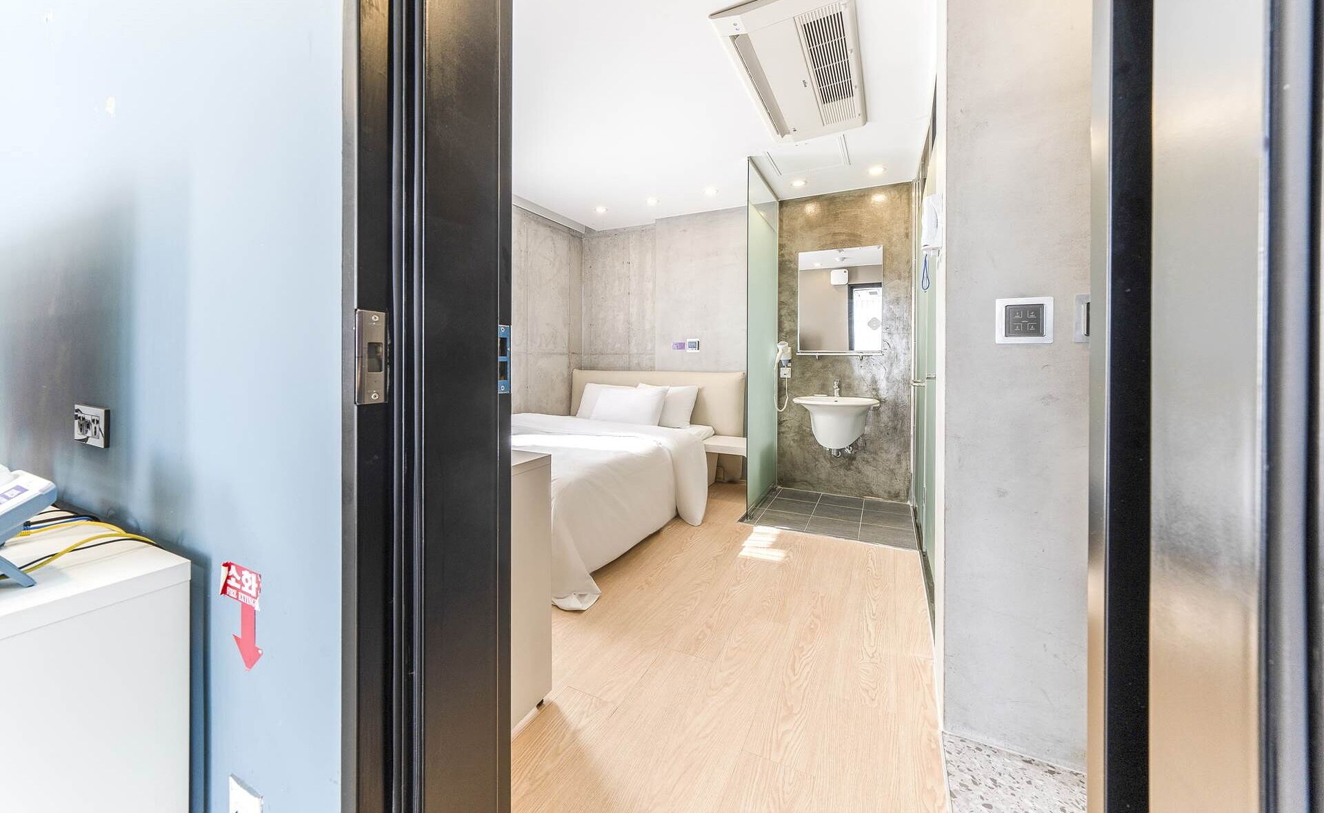 Une chambre d’hôtel avec un lit, une salle de bain dotée d’un lavabo et d’un miroir, et un panneau de commande avec un panneau rouge indiquant la sortie de secours.