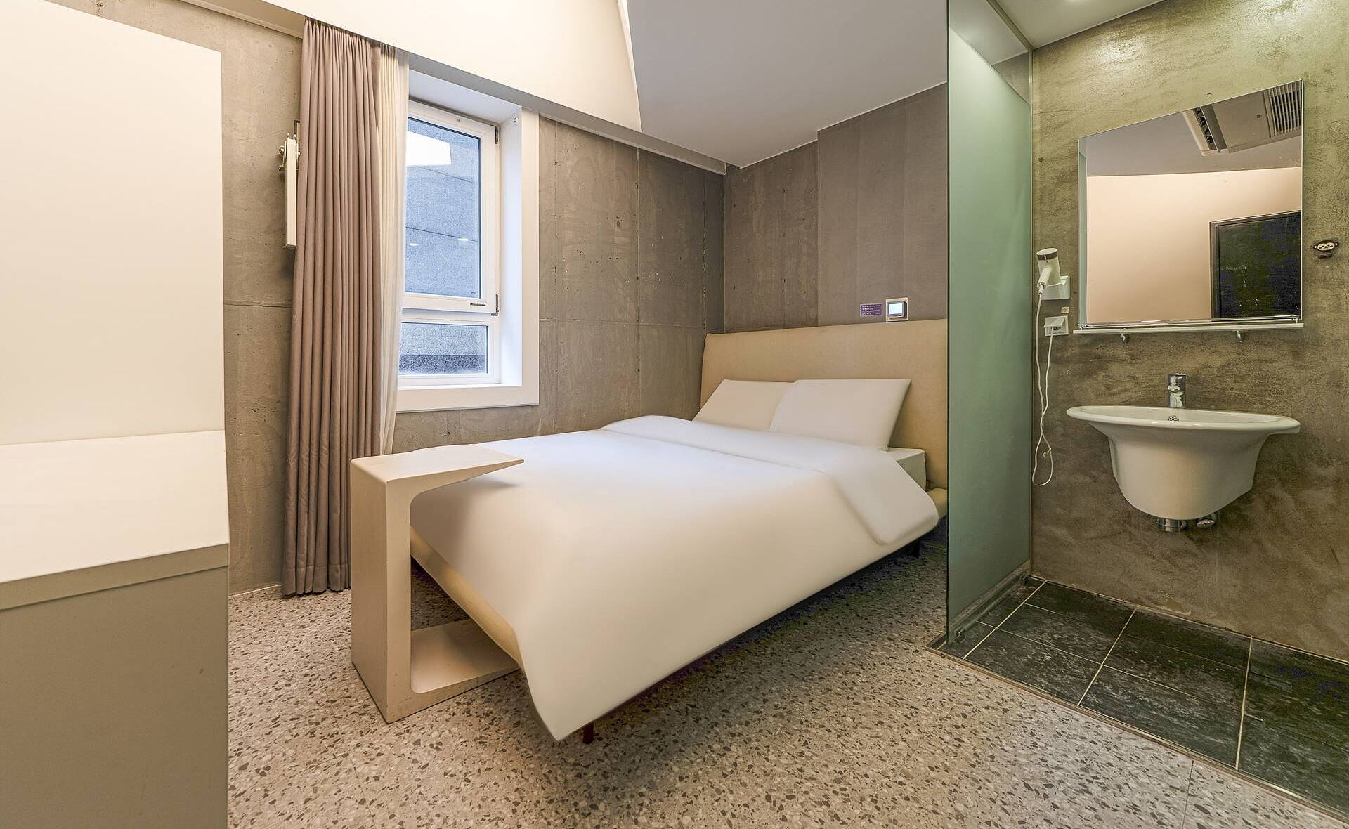 A modern hotel room with a bed, a small bedside table, a window with curtains, a bathroom with a sink and mirror, and a shower area.