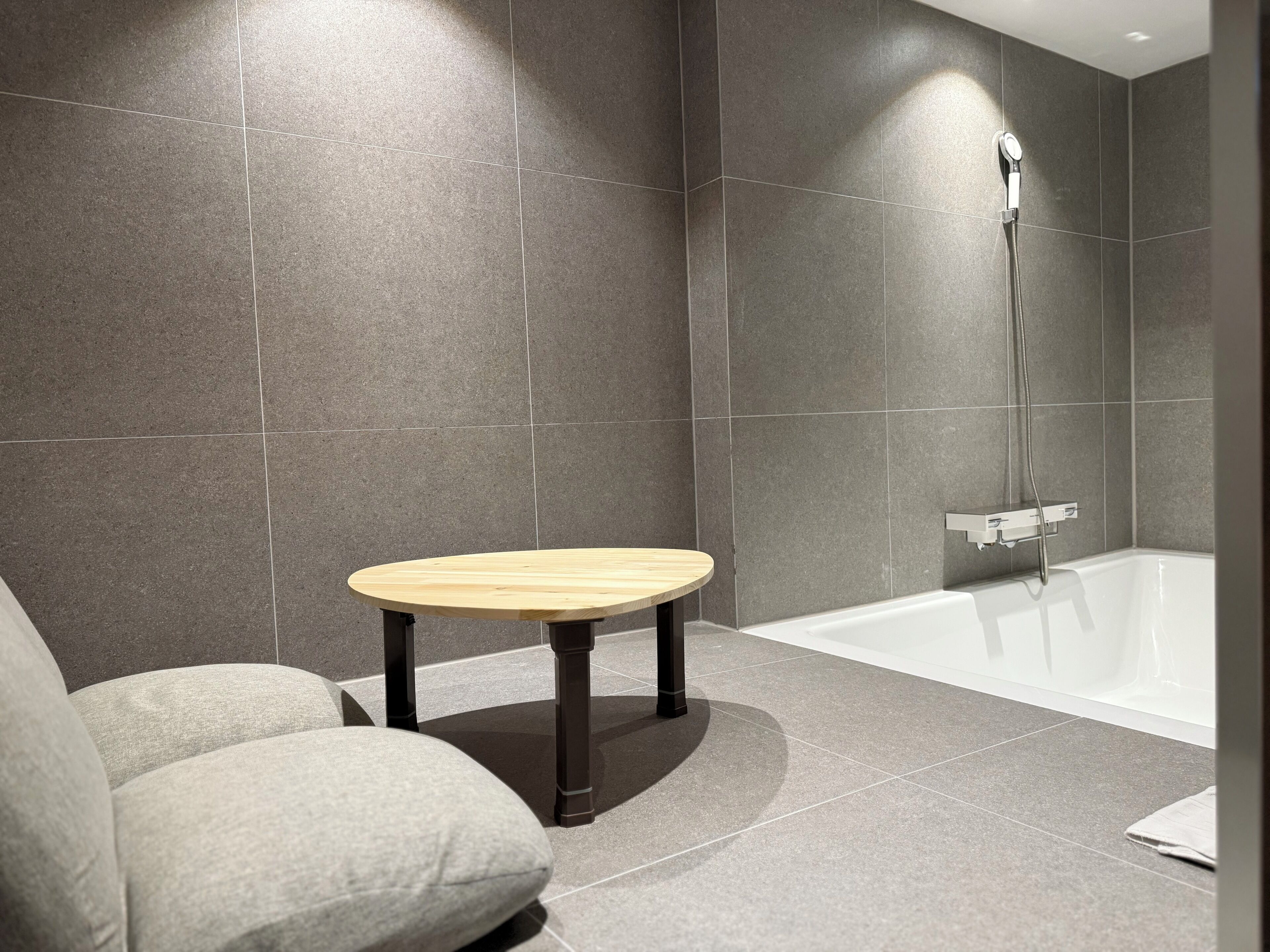 Un baño moderno con bañera, un taburete de madera y paredes con azulejos grises.