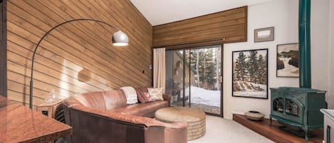 Ein gemütliches Wohnzimmer mit einem braunen Ledersofa, einer Holzwand, einem grünen Kaminofen und einem großen Fenster mit Blick auf Schnee.
