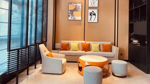 A modern living room with a sofa, armchairs, and ottomans. There are large windows with blinds, artwork on the wall, and a black shelving unit. - The Hotel V Wuxi (Wuxi)