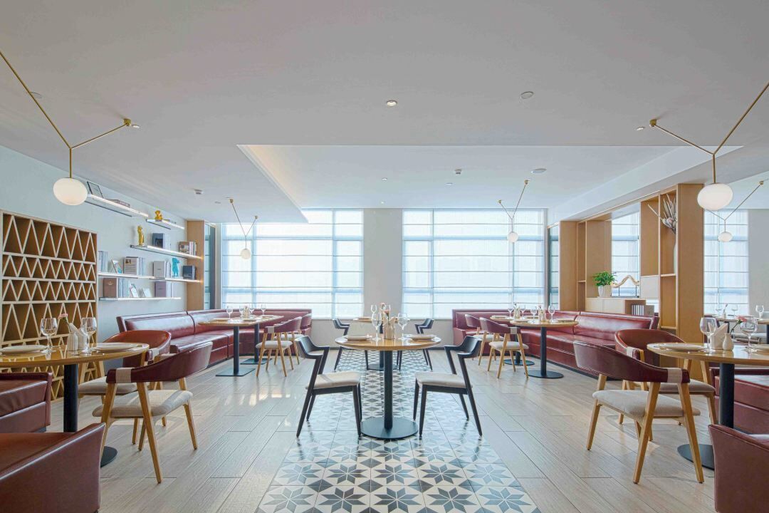 A modern dining area with round tables, red upholstered booths, and a geometric patterned floor.