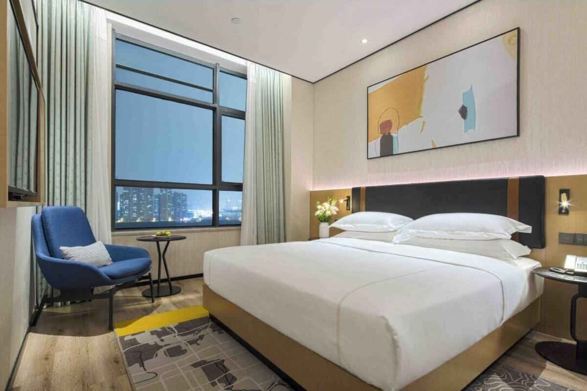 Ein Hotelzimmer mit einem großen Bett, einem Sessel, einem kleinen Tisch und Blick auf die Stadt durch ein großes Fenster.