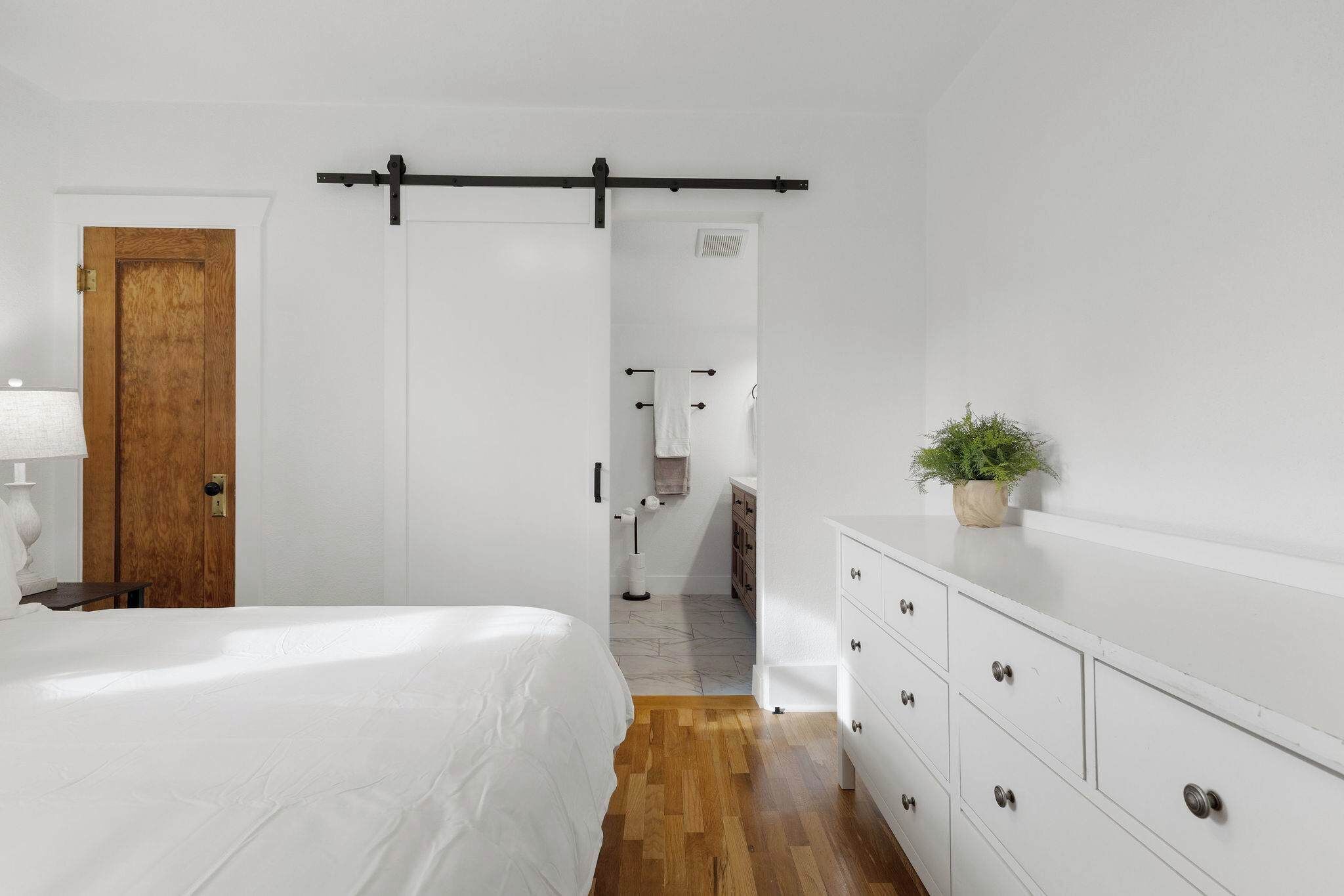 A bedroom with a bed, wooden floor, white walls, a wooden door, a white dresser, and a plant.