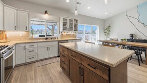 A modern kitchen with white cabinets, a central island, and a dining area with a view. - Harbor House with Hottub on 18th Hole (Klamath Falls)