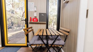 Une pièce avec une table et des chaises en bois, une porte donnant sur la vue d’arbres, une étagère avec un objet rouge, et un objet gris sur le mur.