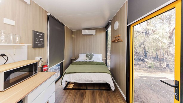 Une pièce de taille modeste comprenant un lit, une petite kitchenette, une fenêtre donnant sur les arbres et une porte menant à l’extérieur.