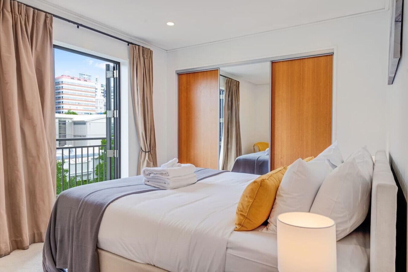 A hotel room with a large bed, a bedside table, a lamp, and a view of the city through the window.