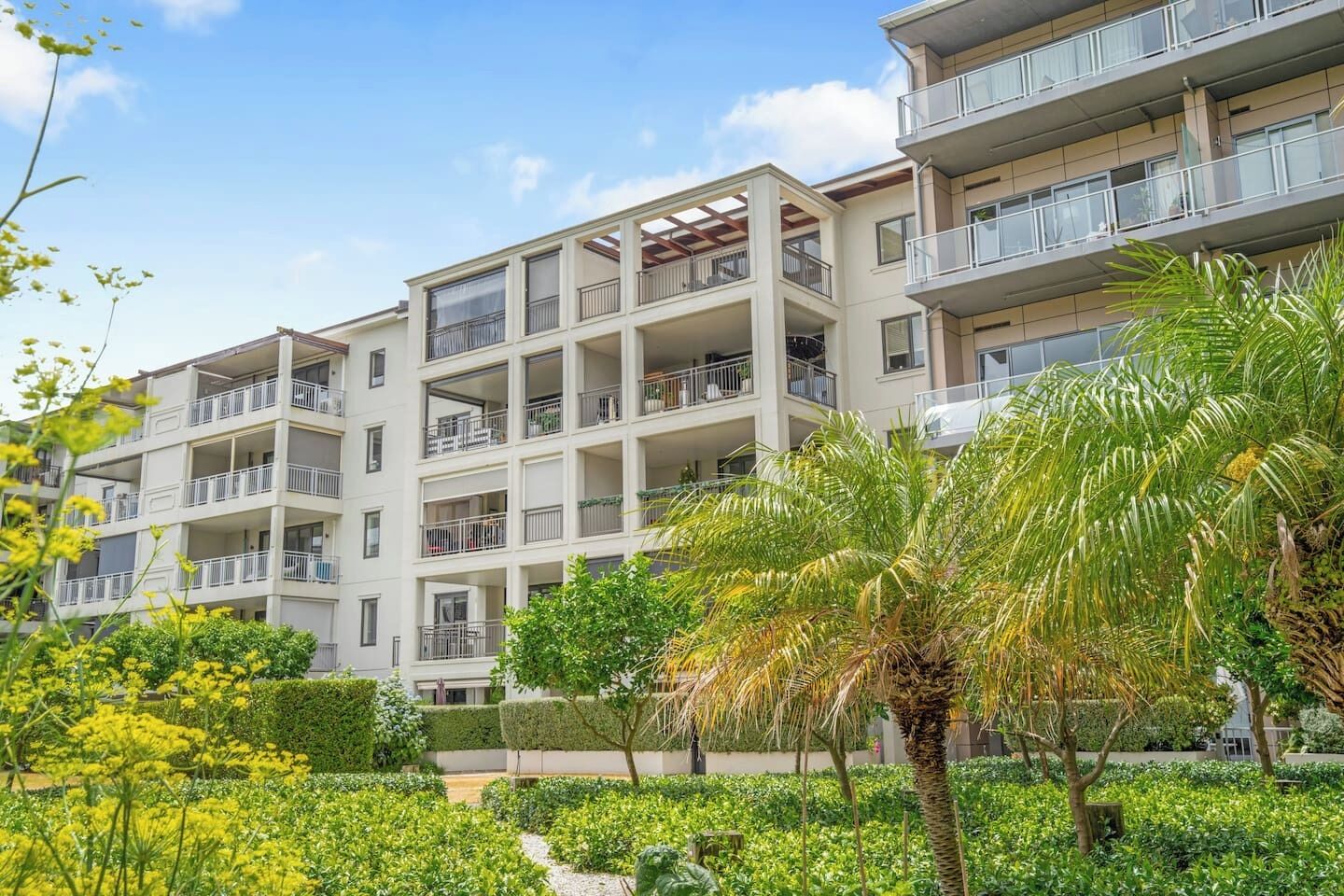 A multi-story residential building with balconies, surrounded by lush greenery and flowering plants.