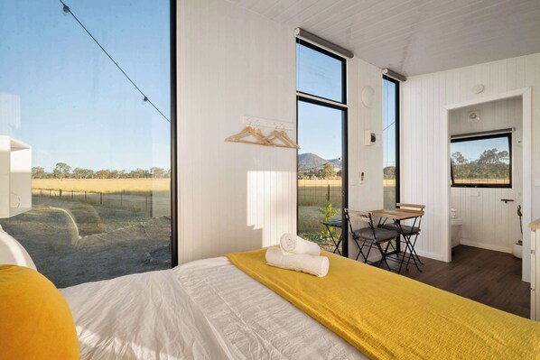 A bedroom with a large bed, a yellow throw, and a view of a field through large windows.